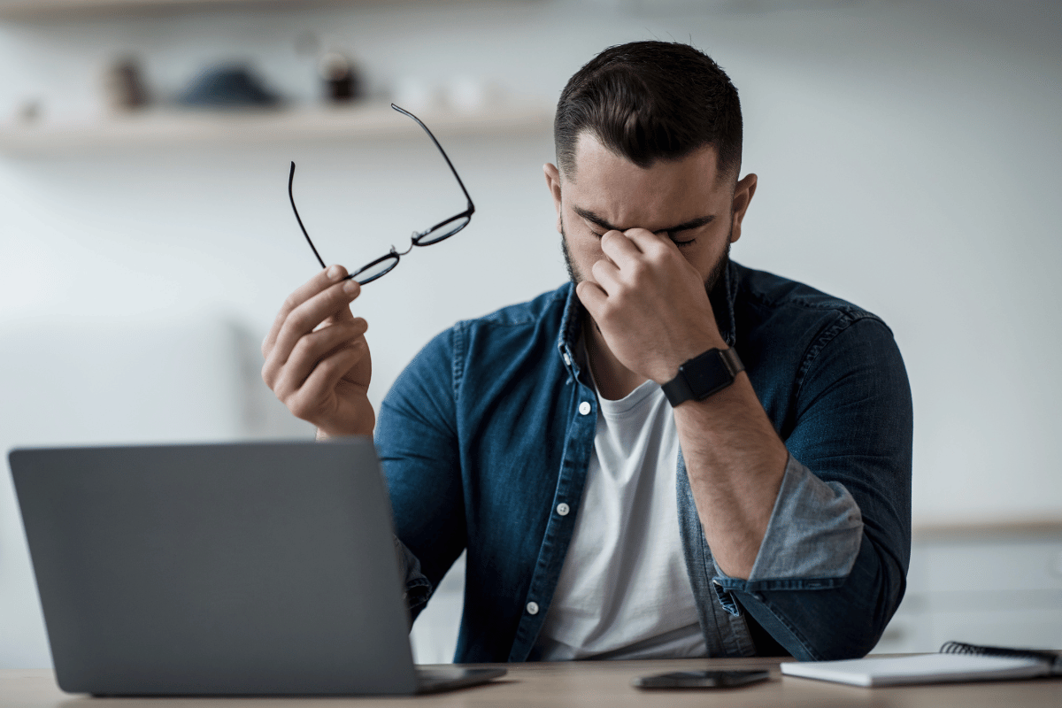 a man sitting in front of his computer, taking off his glasses, and looking uncomfortable